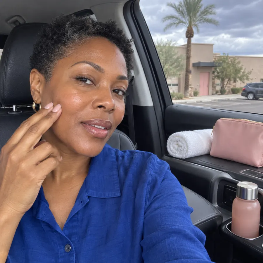 Editorial photo of a Black woman in a cobalt shirt checking her cheek with two fingers in a parked car, with a blush pouch, rolled towel and bottle beside her.